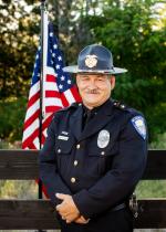 Chief Gibson standing in front of a wooden fence in front of a hanging American Flag, dressed in his Mt. Shasta Police uniform, hands clasped in front of his body, smiling with a grey mustache